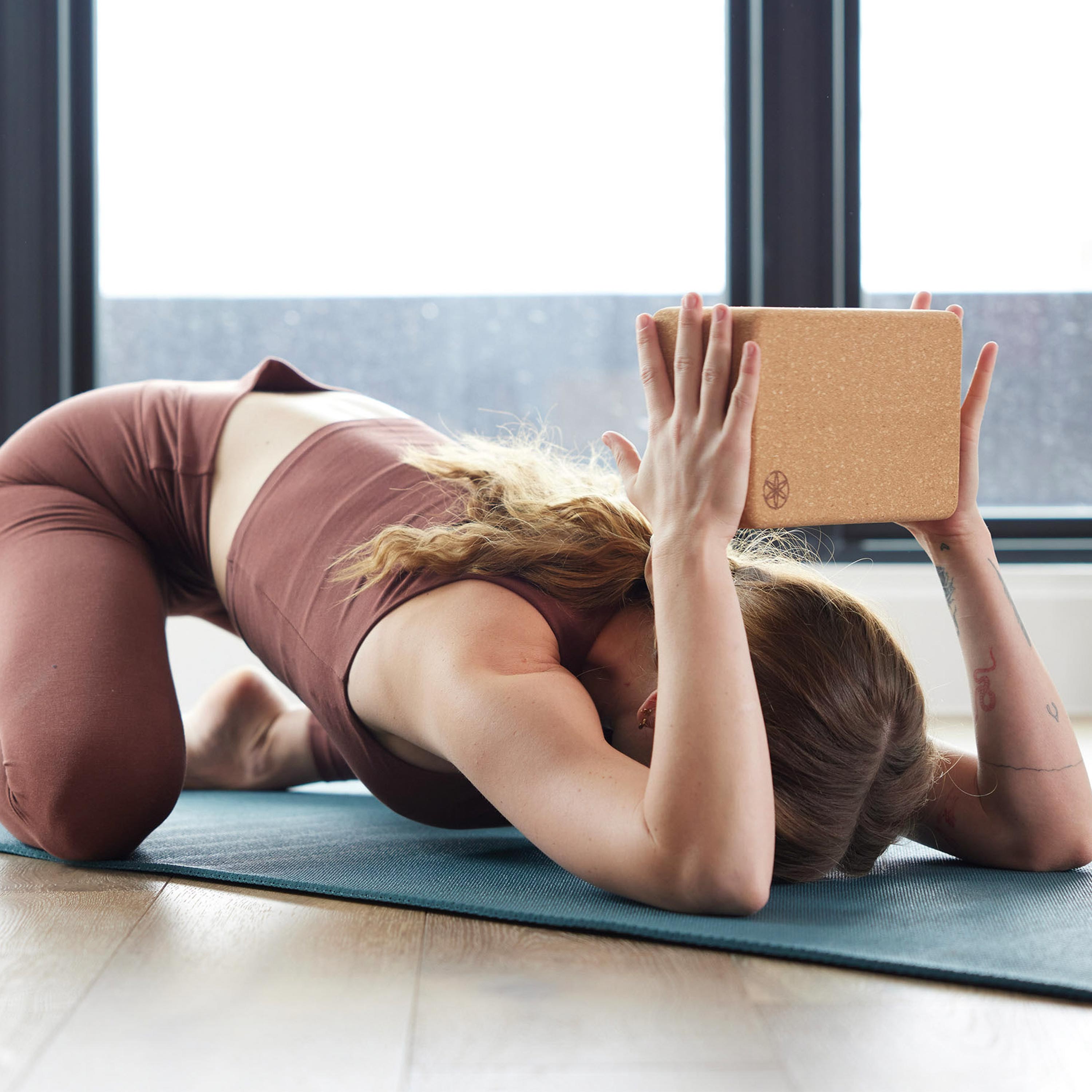 Person on mat in Childs Pose with the Performance Cork Block in hands with arms bent at the elbow - block above back of head.